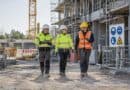 Three construction workers in hard hats and hi-vis walking past scaffolding and a PPE safety sign