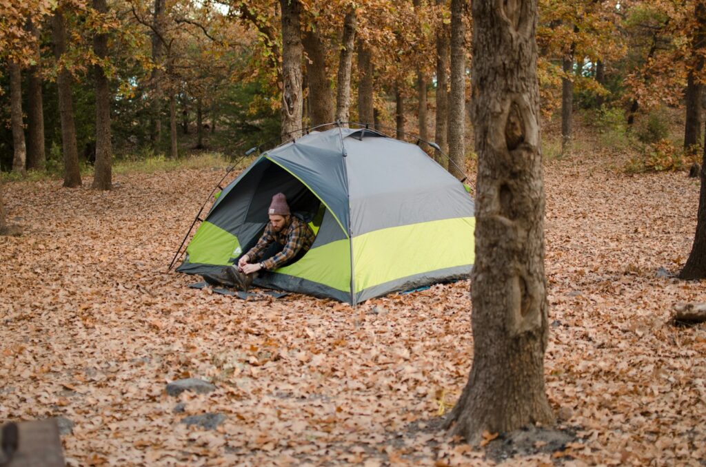 Camper in a beanie and checked shirt lacing his boots in the doorway of a grey and lime dome tent, pitched on fallen oak leaves in autumn woodland