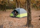 Camper in a beanie and checked shirt lacing his boots in the doorway of a grey and lime dome tent, pitched on fallen oak leaves in autumn woodland