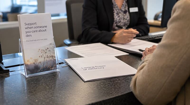 Bank adviser and customer at a counter with a bereavement leaflet and power of attorney papers