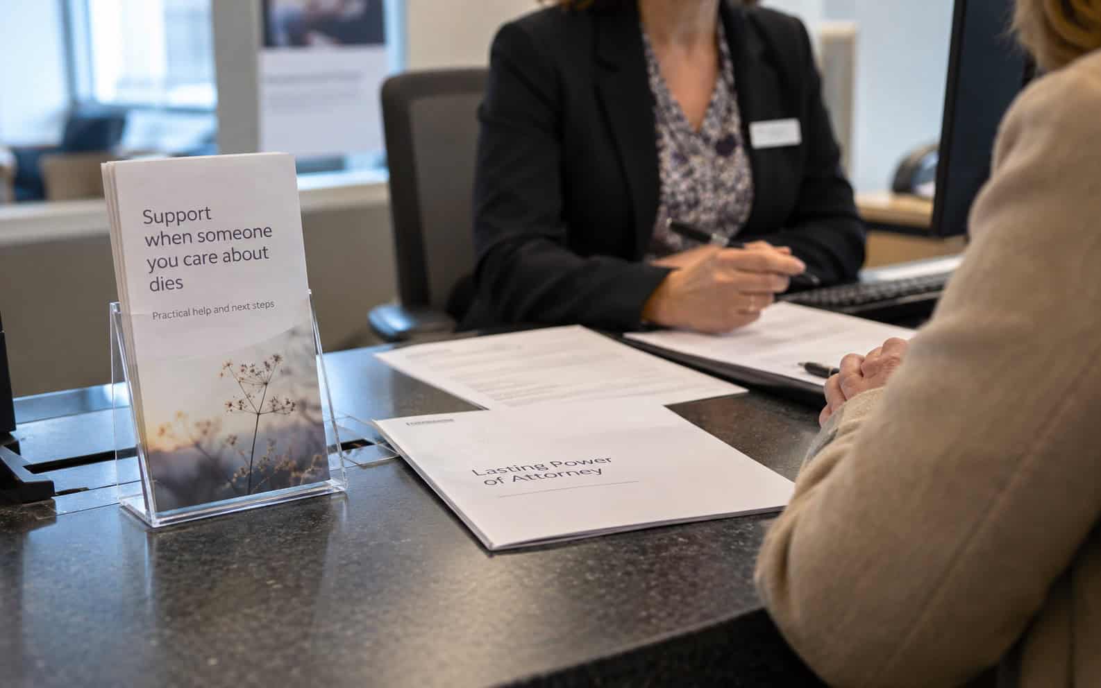 Bank adviser and customer at a counter with a bereavement leaflet and power of attorney papers