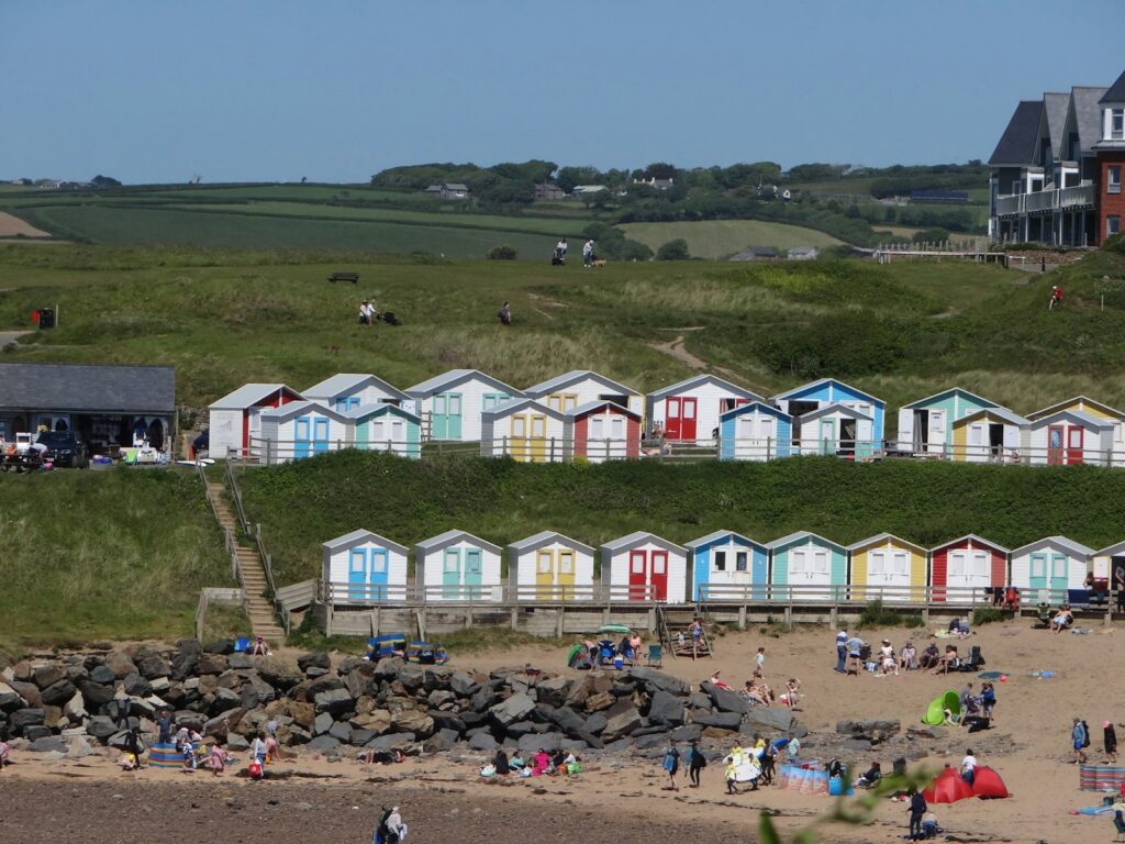Two tiers of colourful beach huts on the grassy slope above a busy sandy beach at Bude, Cornwall
