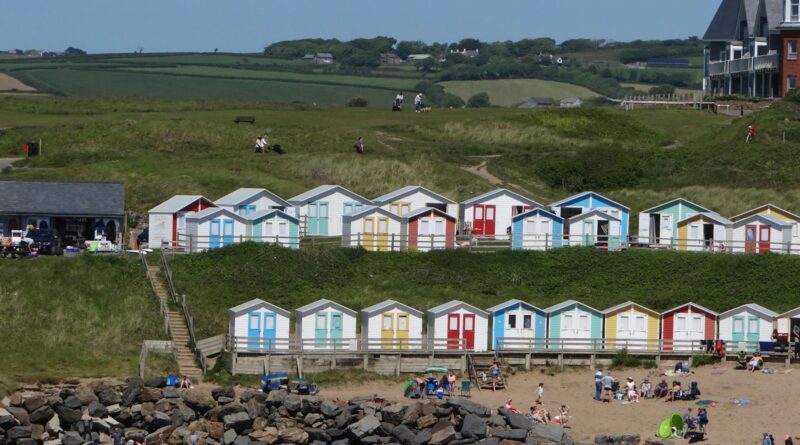 Two tiers of colourful beach huts on the grassy slope above a busy sandy beach at Bude, Cornwall