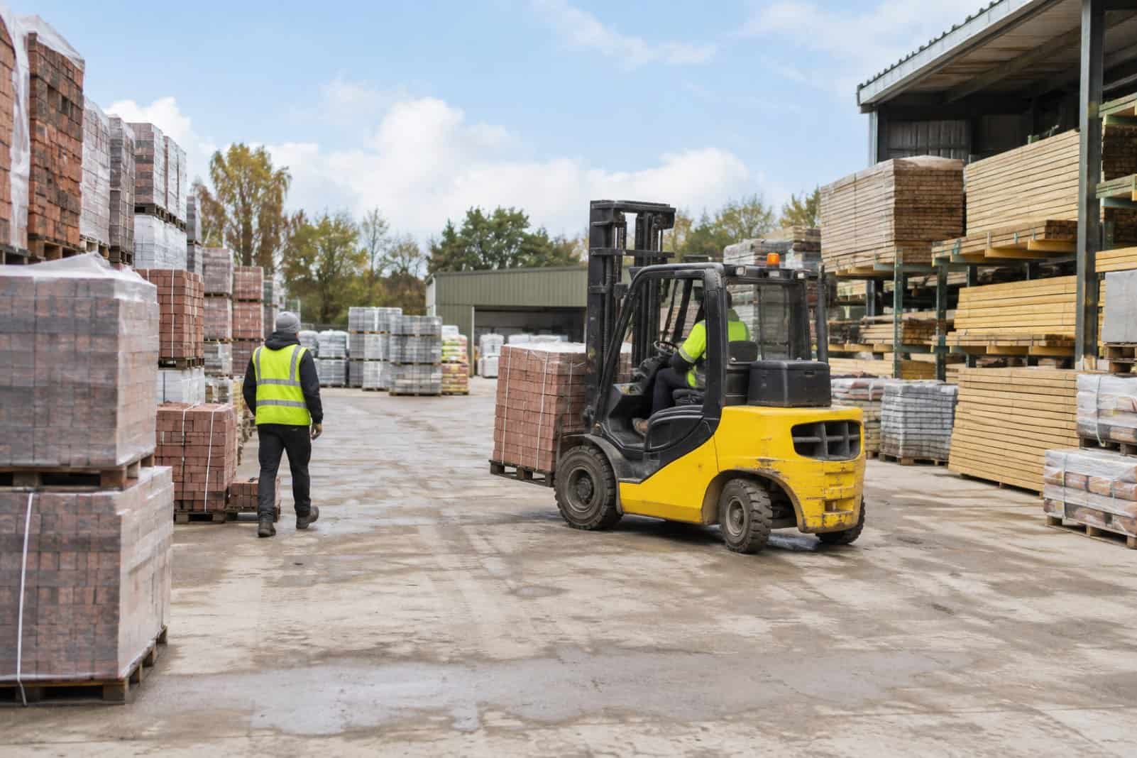 Forklift carrying a pallet of bricks through a builders merchants yard with a worker in hi-vis