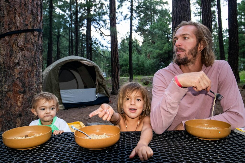 Father and two young children sitting at an outdoor table with bowls of food in a pine forest campsite