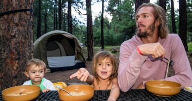 Father and two young children sitting at an outdoor table with bowls of food in a pine forest campsite