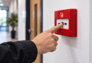 Hand pressing the button on a red fire alarm call point in an office corridor