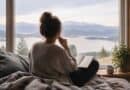 Woman sitting on a bed with a notebook, looking out of a window at a lake and hills