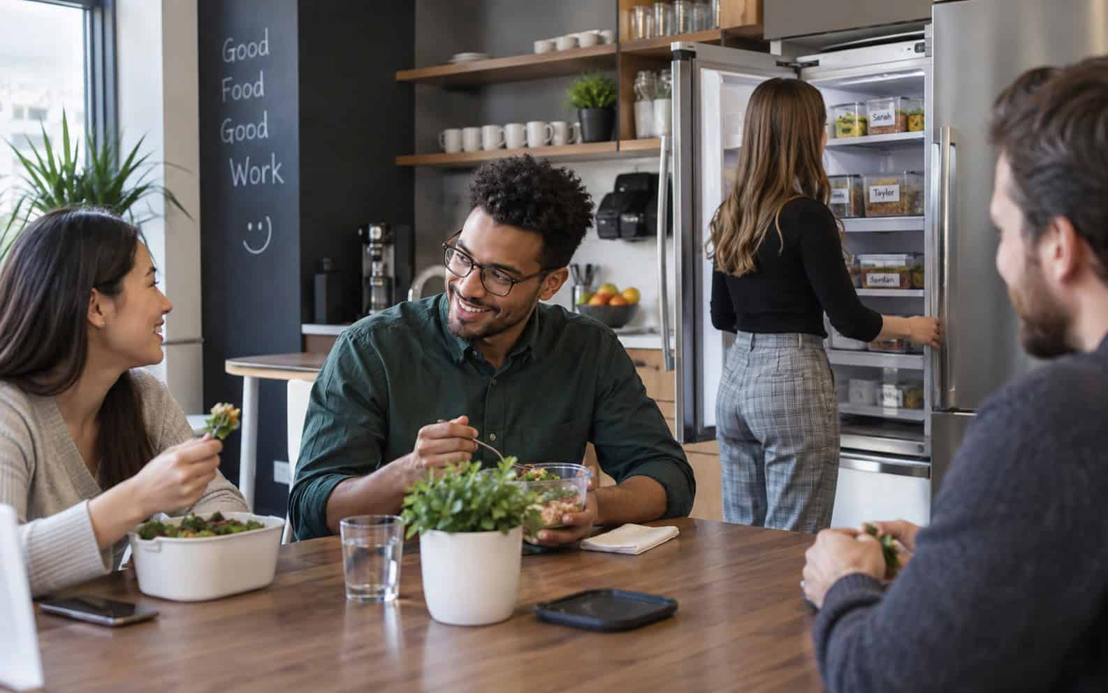 Colleagues eating salads in an office kitchen beside a fridge of name-labelled lunch boxes