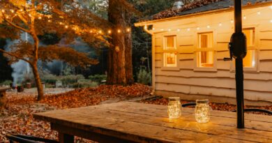 Wooden garden dining table with two glowing jar lanterns, festoon lights and autumn leaves behind at dusk