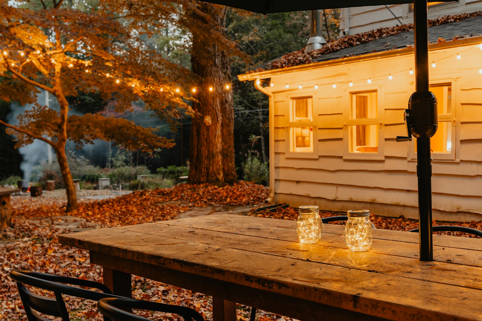 Wooden garden dining table with two glowing jar lanterns, festoon lights and autumn leaves behind at dusk