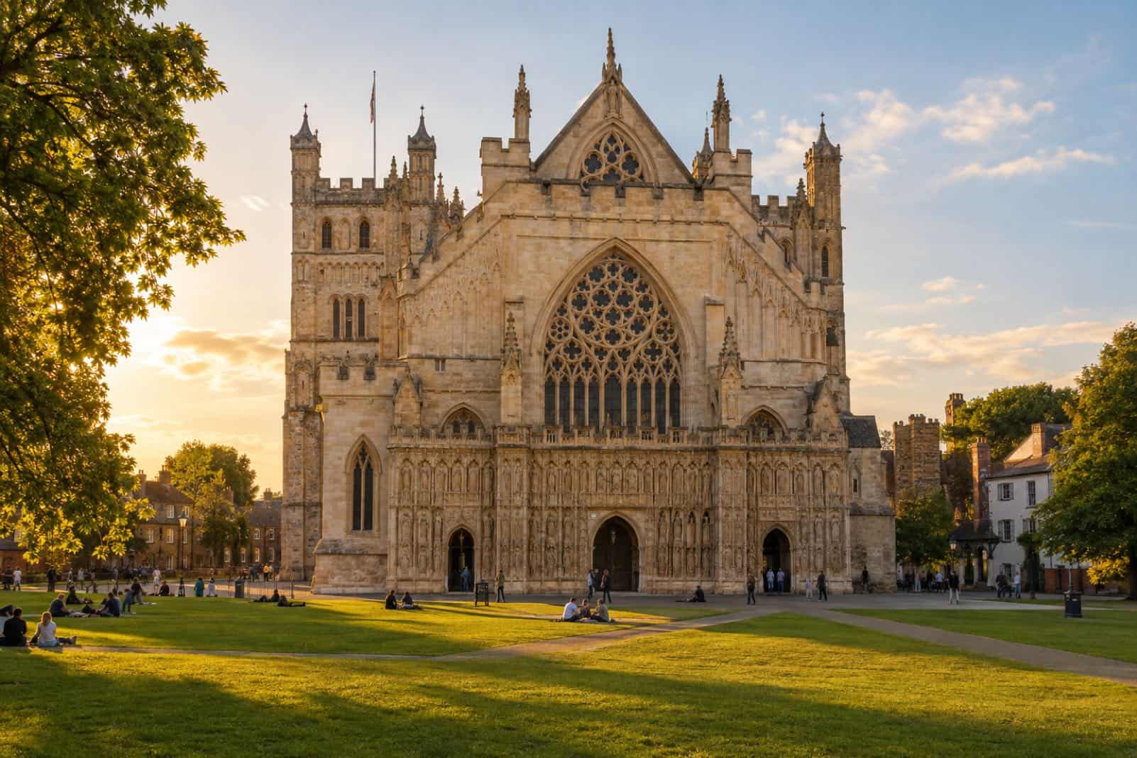 Gothic cathedral west front with a large tracery window in low sunlight, people sitting on the lawn in front