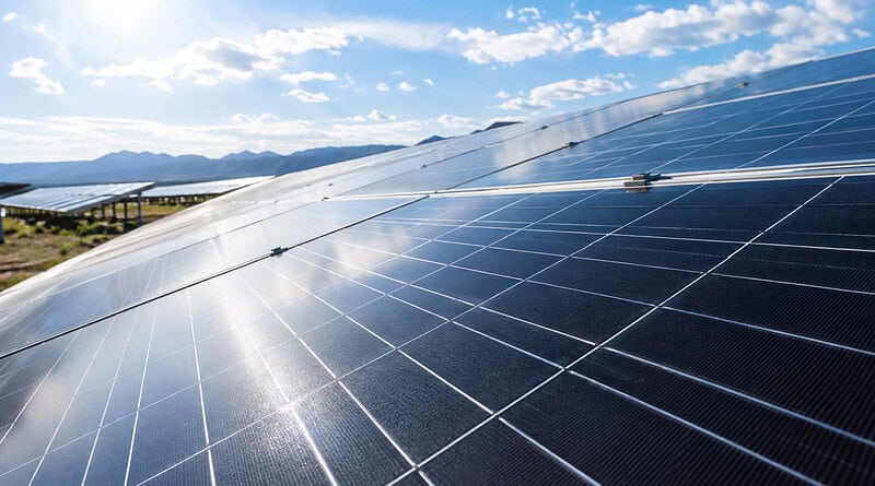 Rows of blue solar panels at a solar farm under a sunny sky with mountains behind