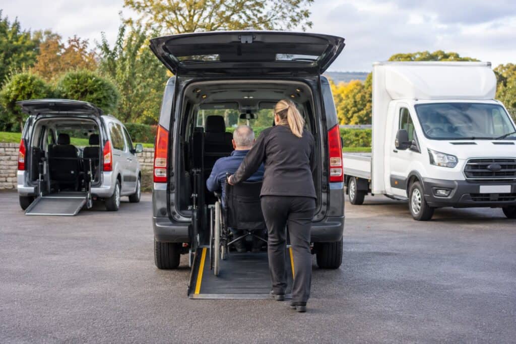 Woman pushing a man in a wheelchair up a rear ramp into a grey accessible vehicle in a car park