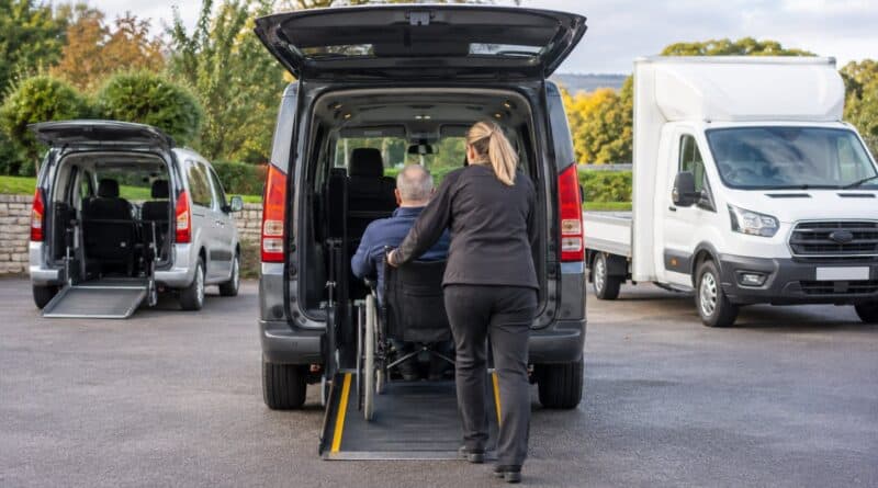 Woman pushing a man in a wheelchair up a rear ramp into a grey accessible vehicle in a car park