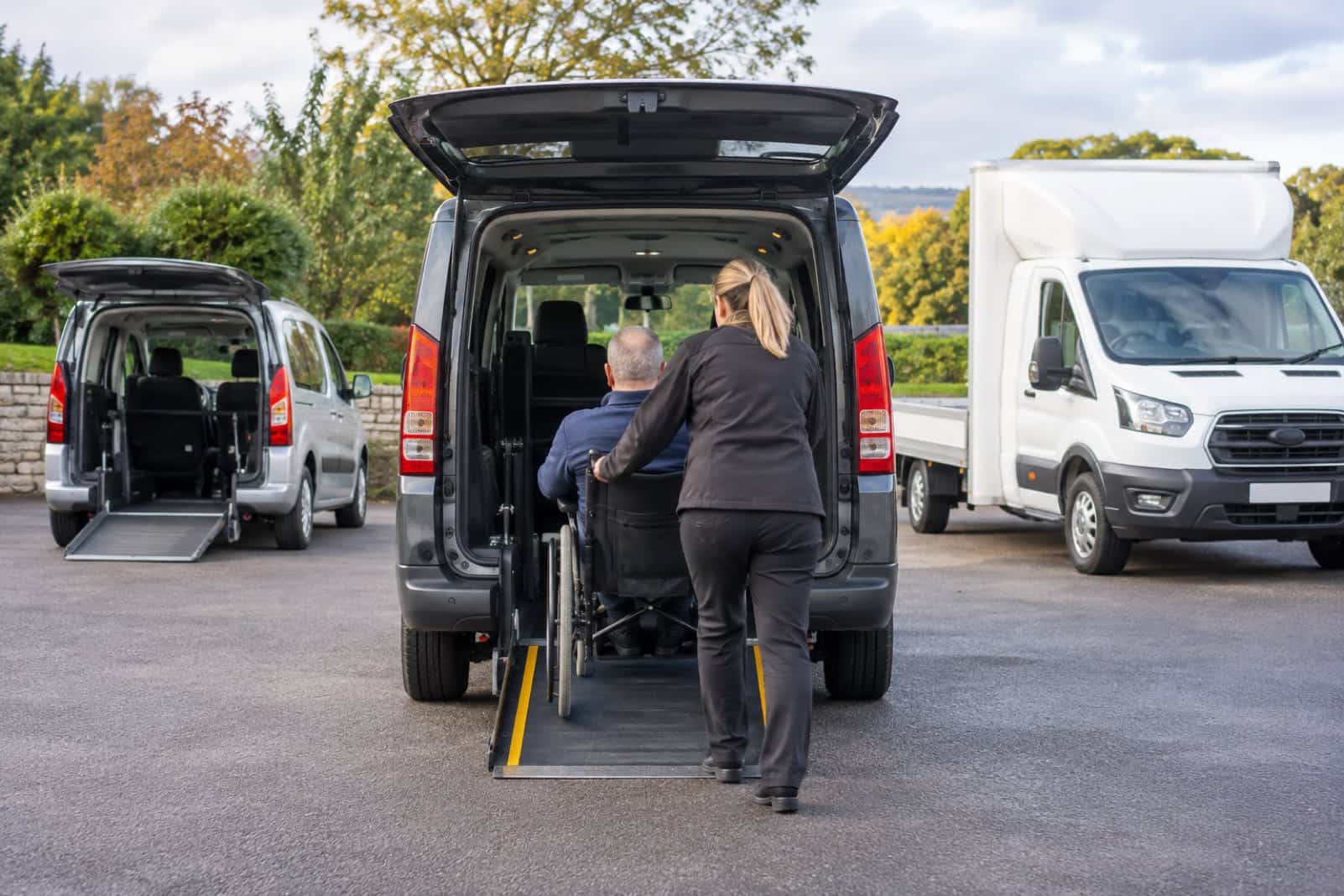 Woman pushing a man in a wheelchair up a rear ramp into a grey accessible vehicle in a car park