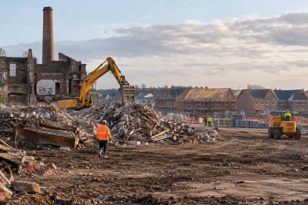 Excavator clearing rubble by a derelict brick mill, with new homes under construction behind