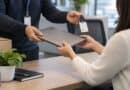 Person handing a closed laptop and a chip card on a lanyard across an office desk to someone in a cream jumper