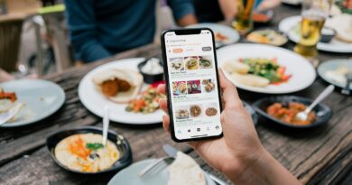 Hand holding a smartphone showing a food menu app above a table of shared dishes at a restaurant