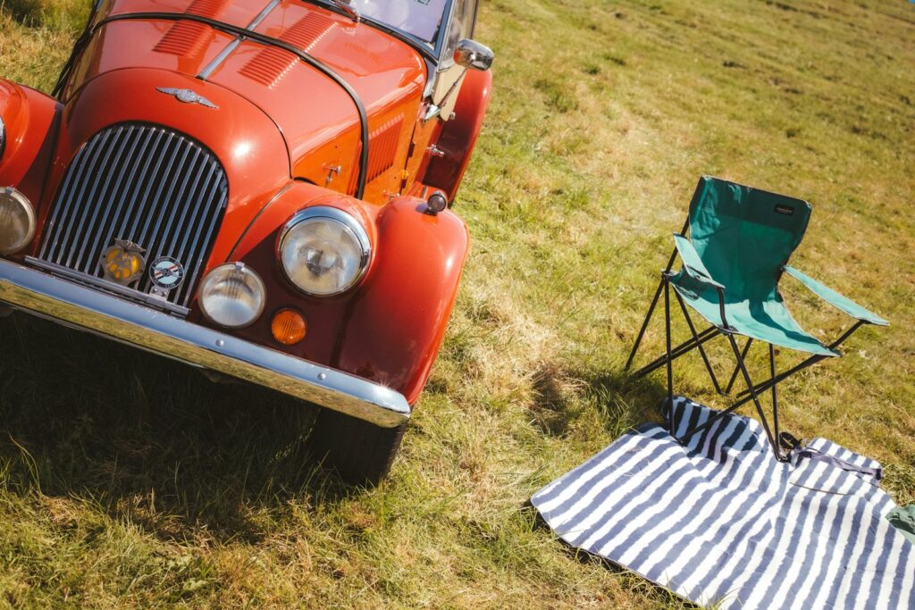 Red classic sports car parked on grass beside a green folding camping chair and a striped picnic blanket