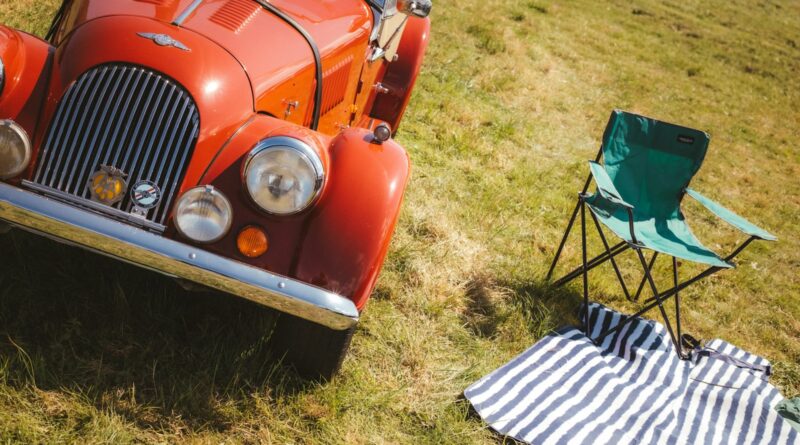 Red classic sports car parked on grass beside a green folding camping chair and a striped picnic blanket