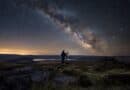 Person with a telescope on moorland at night, a lake below and the Milky Way across the sky
