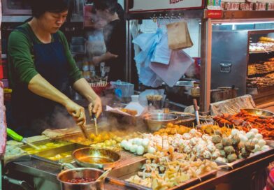 Cook frying skewers and dumplings on a busy street food stall piled with fresh ingredients