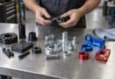 Hands holding a black metal part over a steel bench of black, silver, blue and red machined components