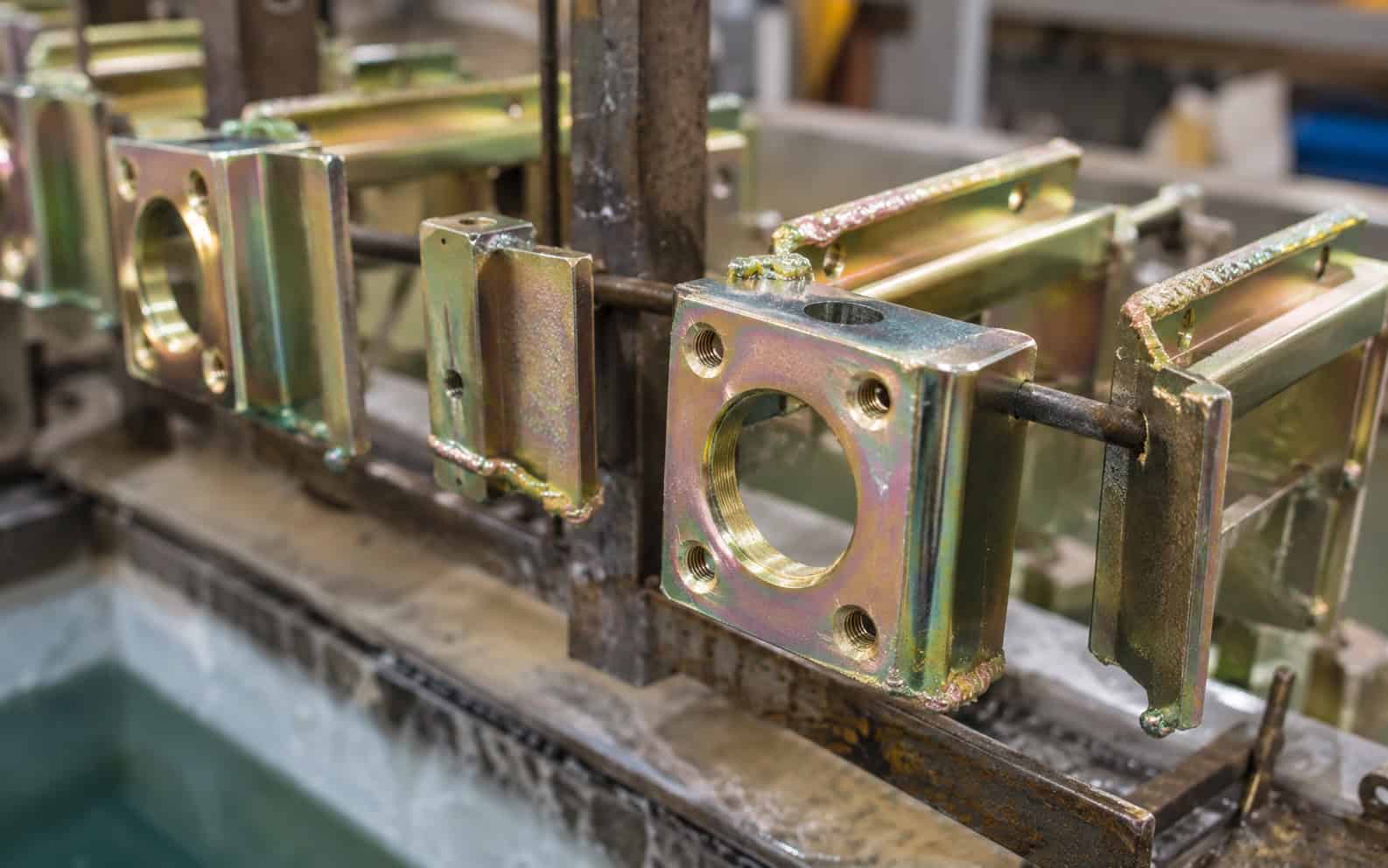 Plated steel brackets with an iridescent gold finish hanging on a jig above a plating tank